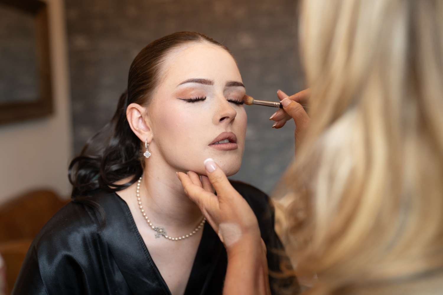 A close-up of a bridesmaid's face as eye makeup is carefully applied — eyes closed, pearl necklace, complete stillness